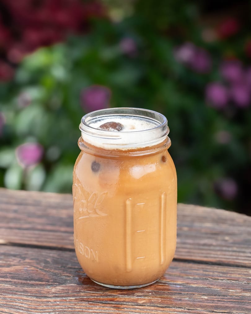 Iced coffee in a mason jar with foam and a chocolate-covered coffee bean on a wooden table.