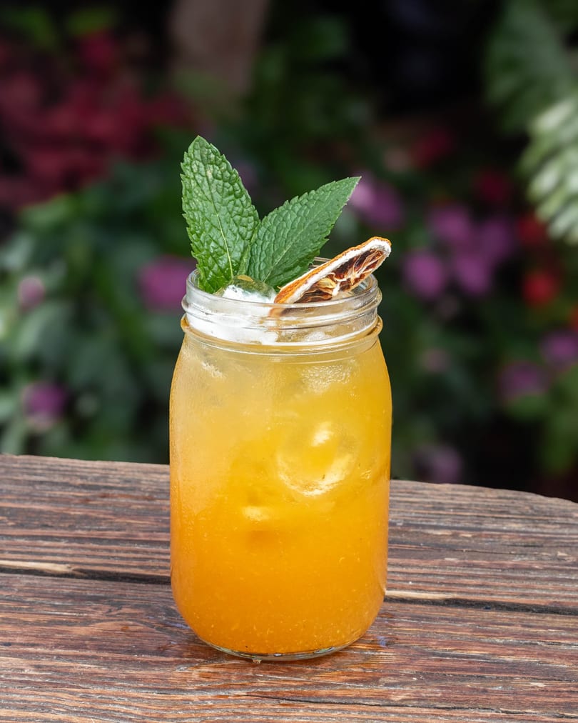 A vibrant orange cocktail in a mason jar, garnished with mint and a dried orange slice, on a wooden table.