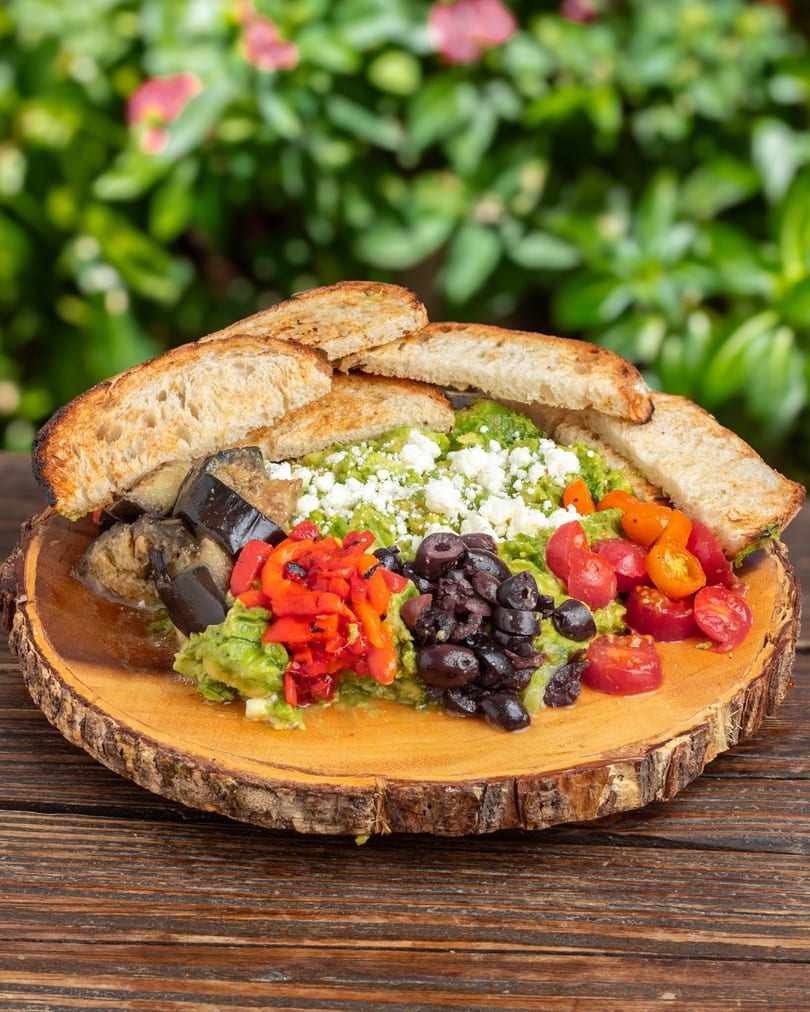 Avocado toast with feta, roasted peppers, eggplant, olives, and tomatoes on a wooden board.