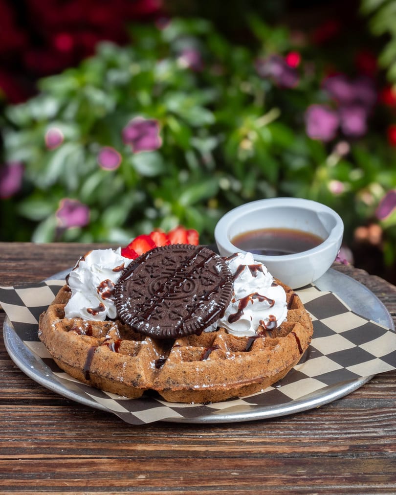 A waffle topped with whipped cream, chocolate syrup, an Oreo cookie, and sliced strawberries.