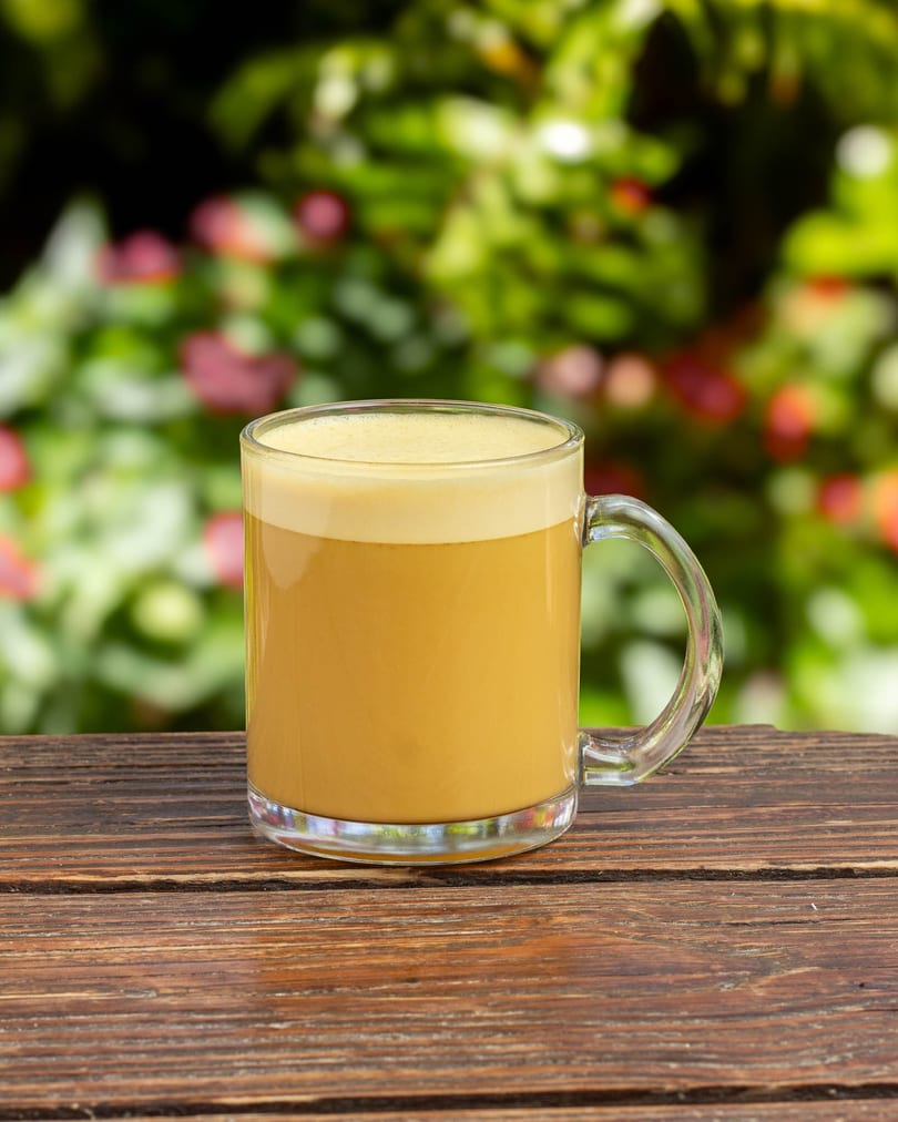 A clear mug of golden frothy beverage sits on a wooden table with a blurred green and pink background.