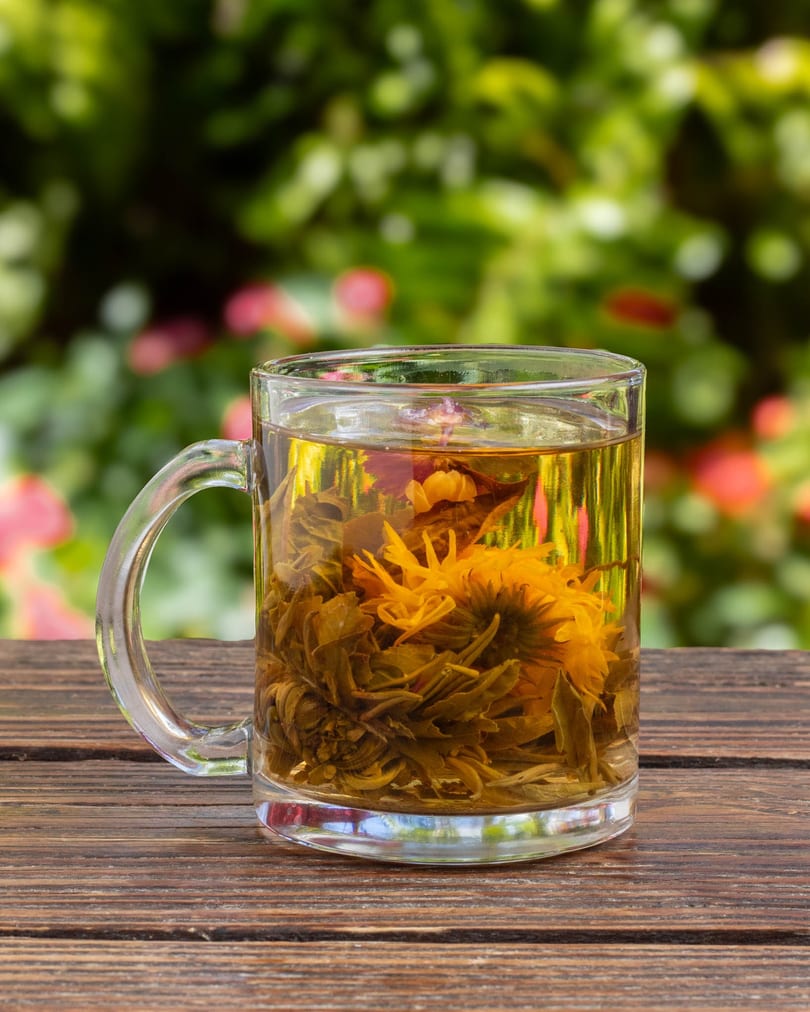 Blooming tea in a clear glass mug on a wooden table with a blurred green and pink background.