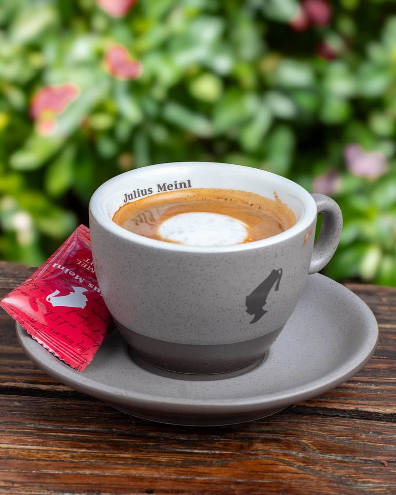 A Julius Meinl coffee cup with foam and a red sugar packet on a wooden table outdoors.