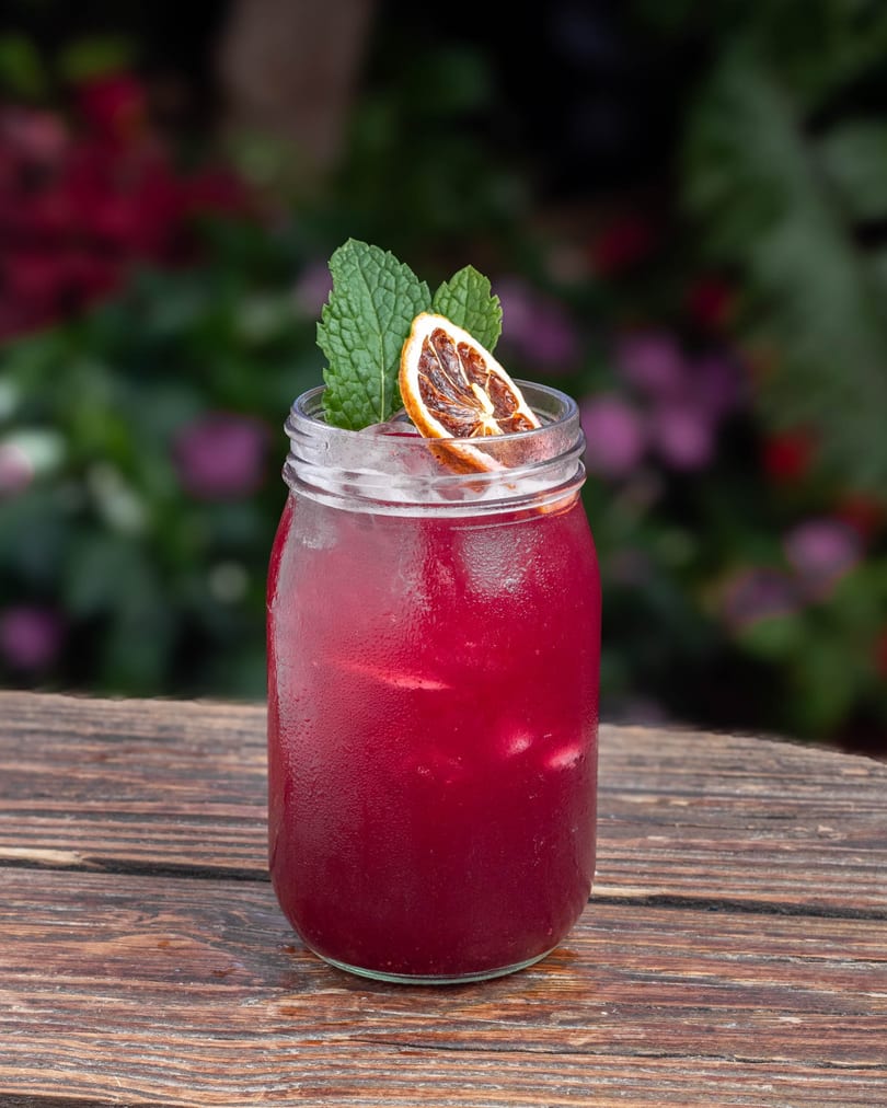 A vibrant red drink in a mason jar, garnished with mint and a dried orange slice, on a wooden table.