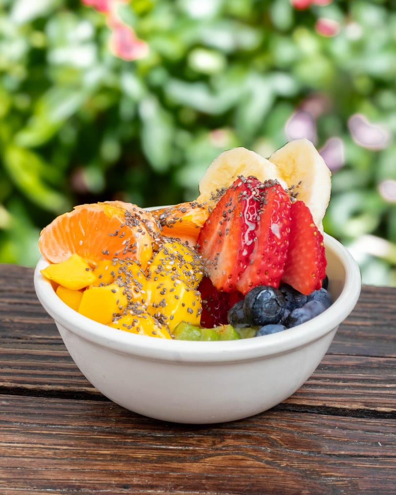 A white bowl filled with fresh fruit, including mango, strawberries, blueberries, and bananas, on a wooden table.
