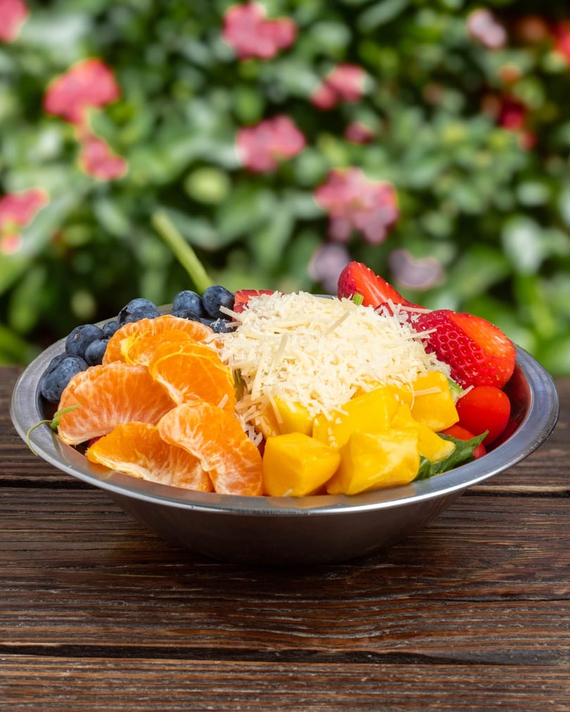 A colorful fruit salad with blueberries, tangerines, mango, strawberries, and shredded cheese in a metal bowl.