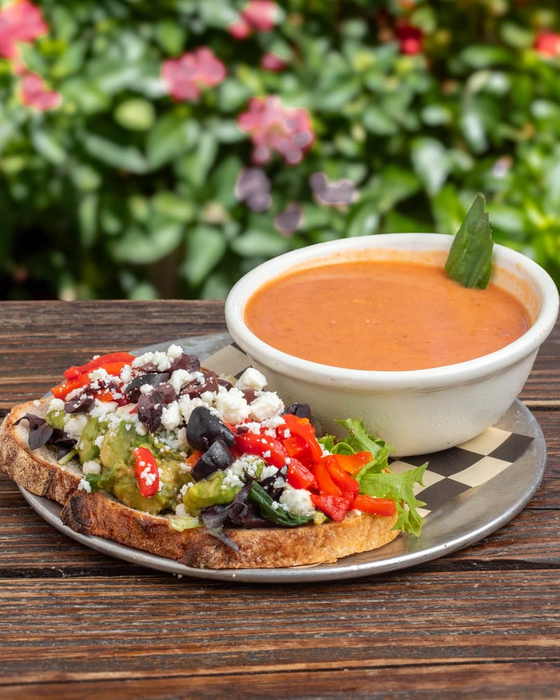 Avocado toast with feta, olives, and red peppers, served with a bowl of tomato soup.