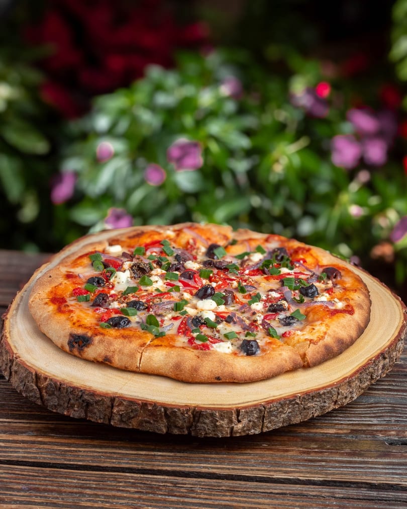 A freshly baked pizza with olives, red peppers, feta, and green onions on a wooden board.