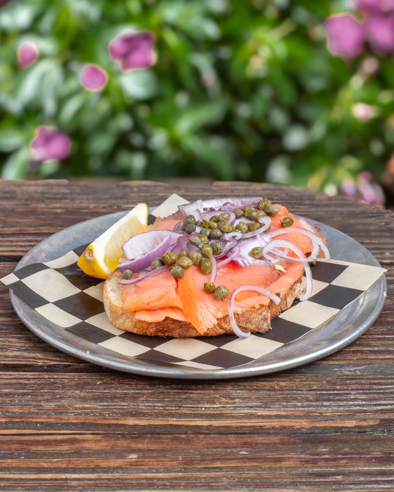 Smoked salmon toast with red onions, capers, and a lemon wedge on a metal plate.