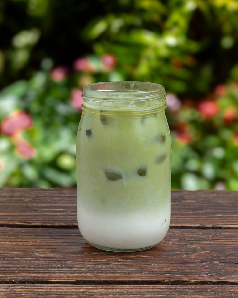 Iced matcha latte in a mason jar on a wooden table with a blurred green and pink floral background.